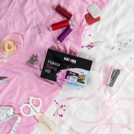 Kadam Pakka Rang Kit placed on a pink fabric with sewing essentials like scissors, threads, ribbons, and pins – perfect fabric dye kit for cotton, silk, wool, and georgette.
