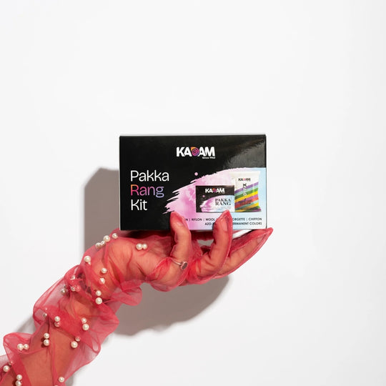 Hand in a stylish red net sleeve with pearls holding Kadam Pakka Rang Kit box – premium fabric dye kit for vibrant permanent colors.