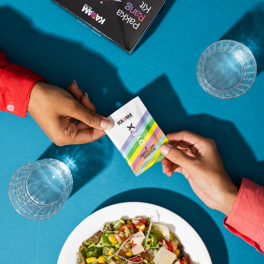 One hand passing Kadam Colors DyFix sachet to another over a vibrant lunch table, symbolizing daily fabric care.