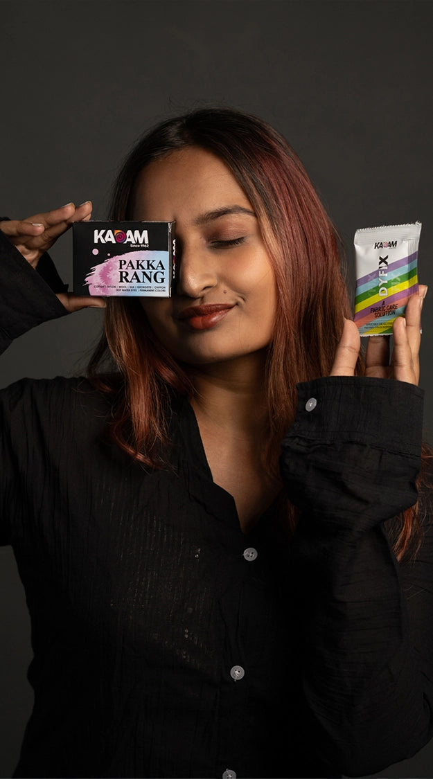 A woman holding Kadam Pakka Rang fabric dye and Dyfix color fixer sachets with a confident smile against a dark background, showcasing vibrant and durable fabric dyeing solutions