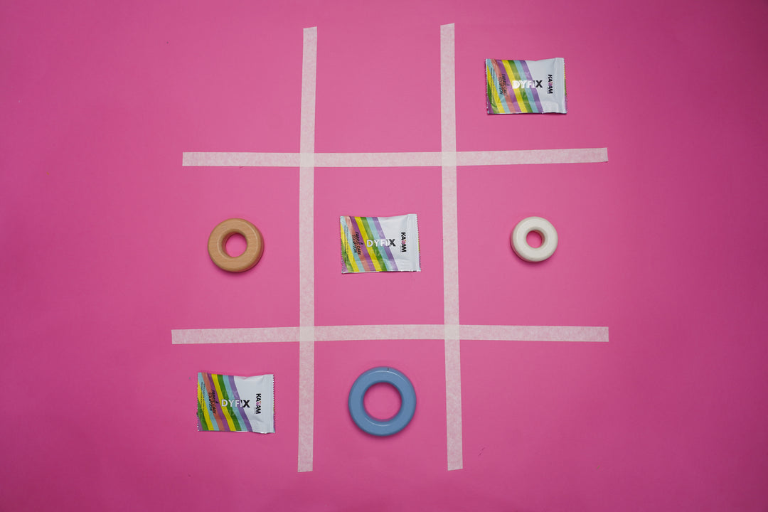 Creative tic-tac-toe game concept using Kadam DyFix sachets on a pink background—fabric care with a fun twist.