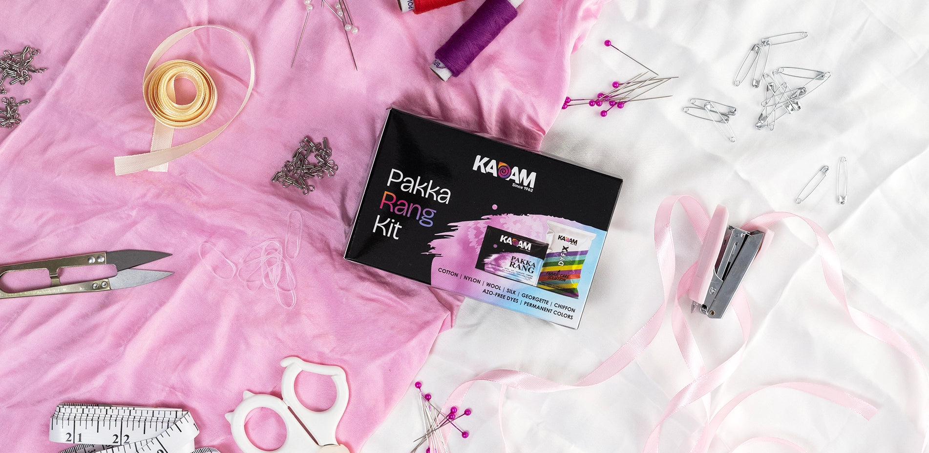 Kadam Pakka Rang fabric dye kit placed on pink fabric with sewing tools, permanent azo-free dye for cotton, silk, wool and DIY clothing projects.