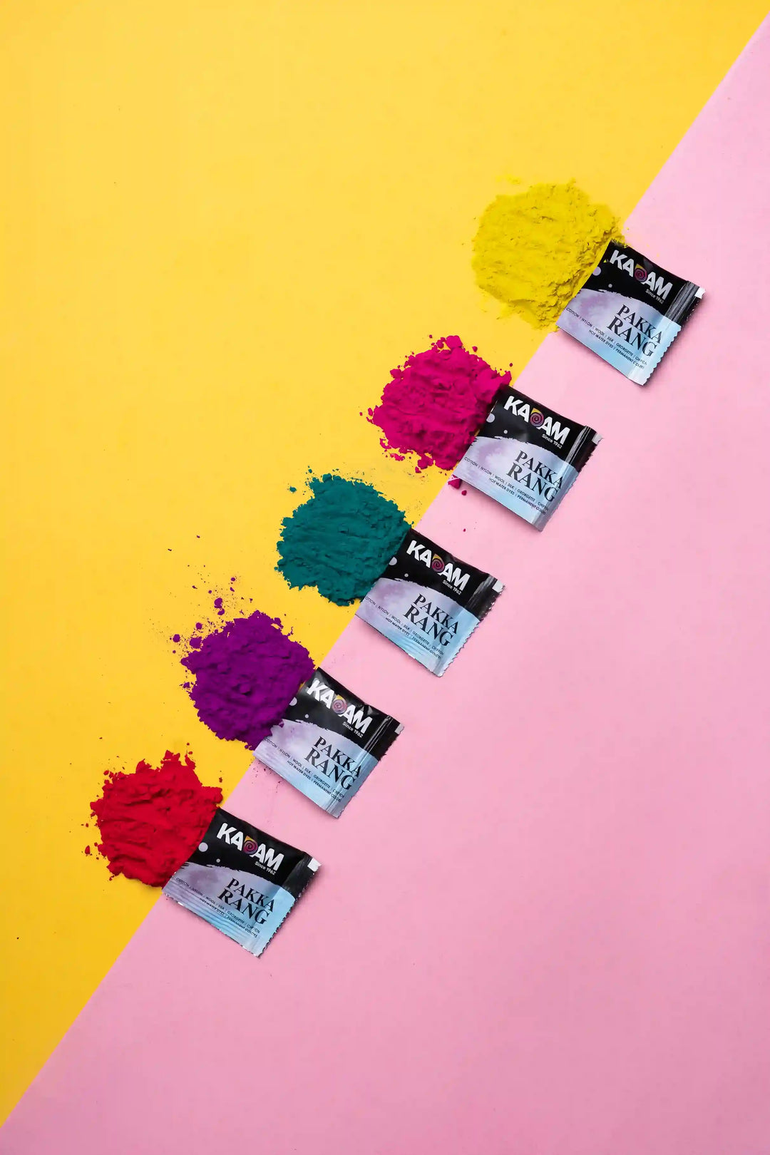 Vibrant Kadam Colors dye powders spilled from sachets on a half yellow, half pink flat lay.