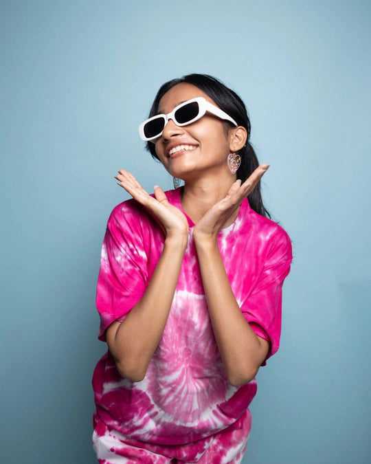 Smiling young woman wearing white sunglasses and a vibrant pink tie-dye t-shirt, posing joyfully against a light blue background.