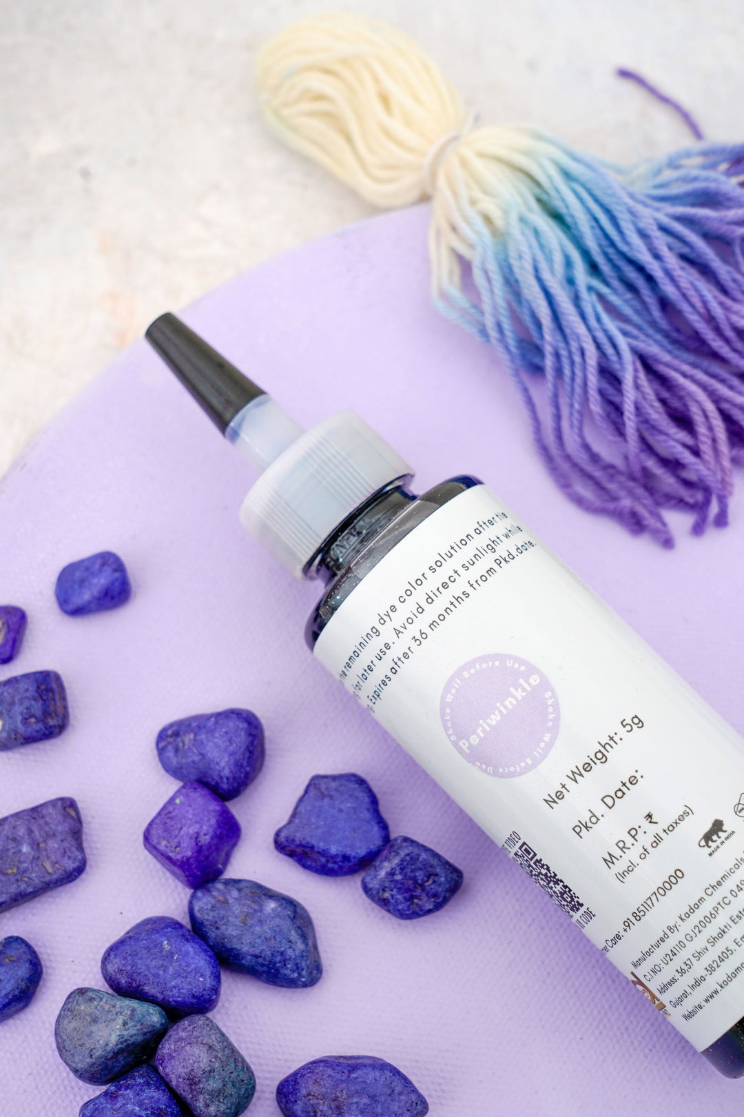 A bottle of Kadam Colors dye placed on a light purple surface, surrounded by vibrant blue dye stones and a gradient-dyed yarn bundle, showcasing the tools and materials for creative dyeing projects