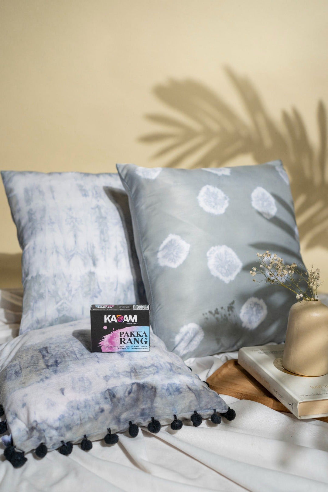 A cozy arrangement of tie-dyed cushion covers in soft gray tones with unique patterns, created using Kadam Pakka Rang fabric dye, displayed alongside a vase with dried flowers and books, showcasing stylish and creative home decor