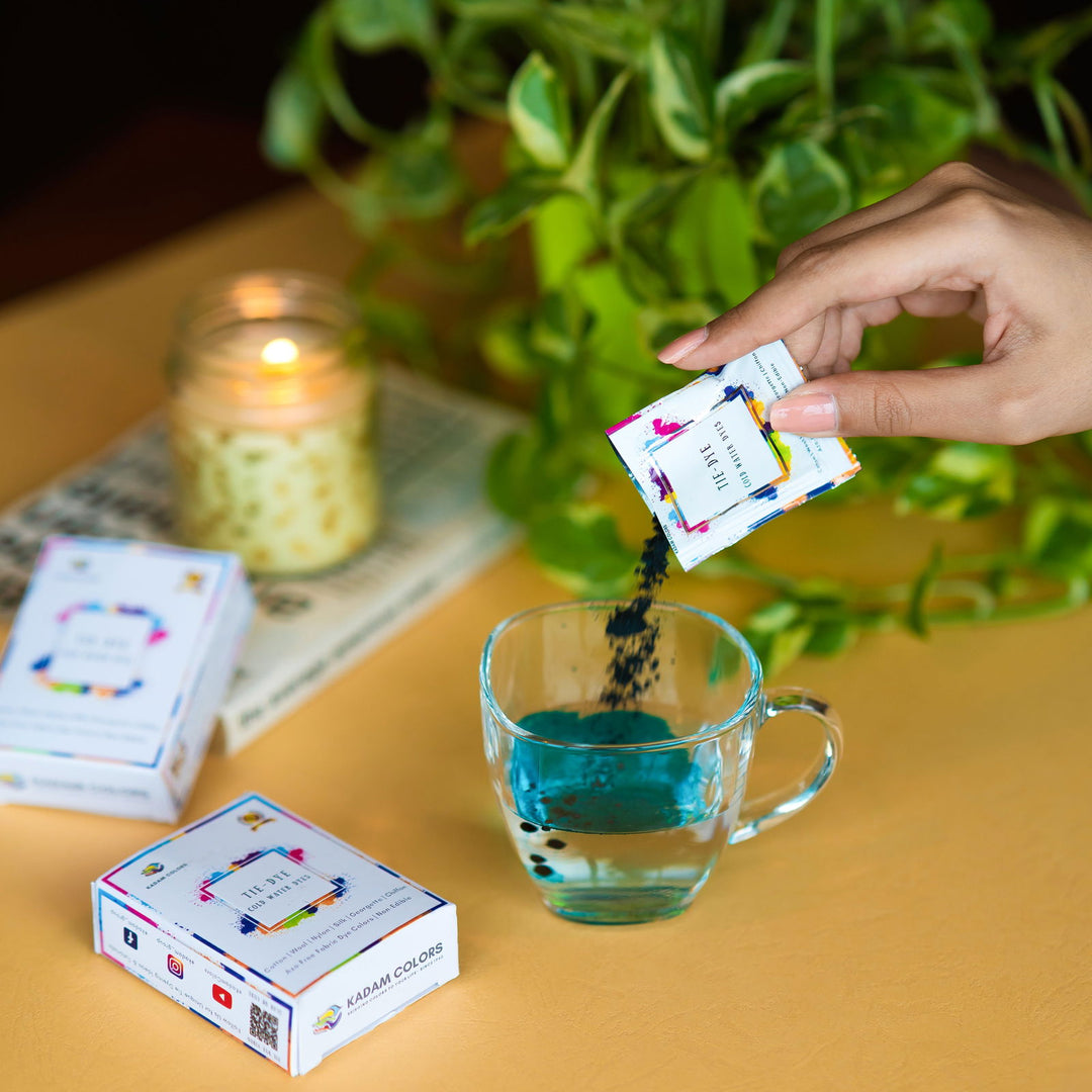 A hand pouring vibrant blue Kadam Colors dye into a glass of water, with the dye boxes and a lit candle in the background, surrounded by green plants, showcasing the ease of use and creative potential of the dye