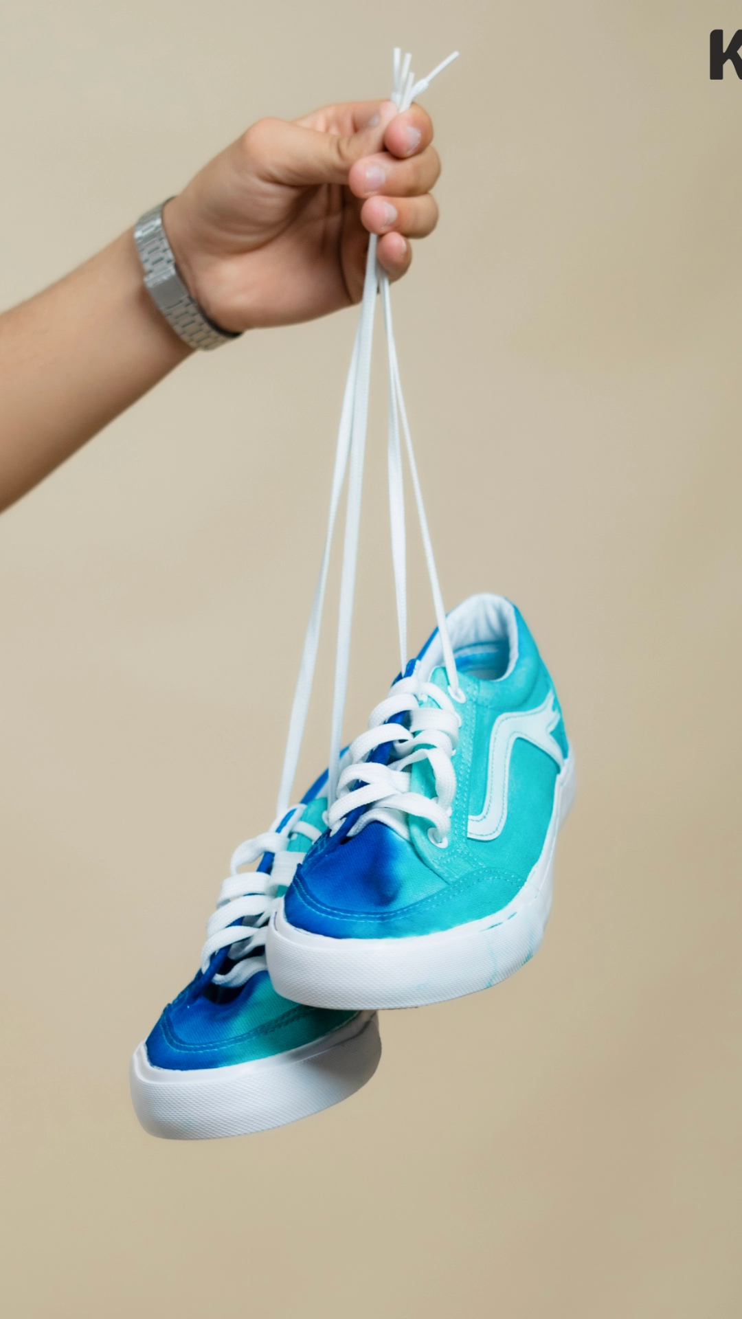 A pair of vibrant blue and teal tie-dyed sneakers with white laces, held by a hand wearing a silver watch, displayed against a beige background, showcasing a creative application of Kadam Colors dyes.