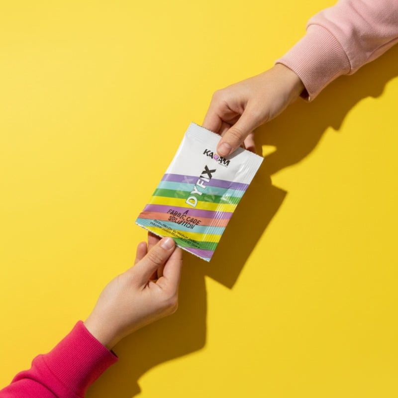 Close-up of two hands exchanging a Kadam DyFix sachet on a bright yellow background, emphasizing easy-to-use single-use packaging.